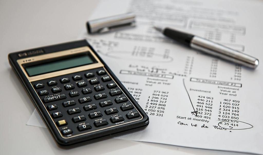 A calculator, a silver pen, and printed financial documents on a table, displaying numerical data and notes.
